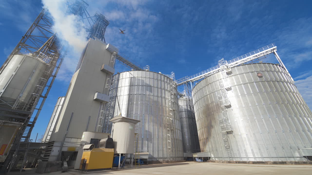Panoramic view of agricultural plant. Large round grain elevators for storing grains. Thick fumes releasing into the air during grain processing on a factory.