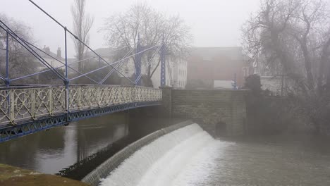 Ornate-Victorian-iron-pedestrian-bridge-over-a-weir-on-the-river-Leam-on-a-cold-misty-day-in-Leamington-Spa,-Warwickshire,-England