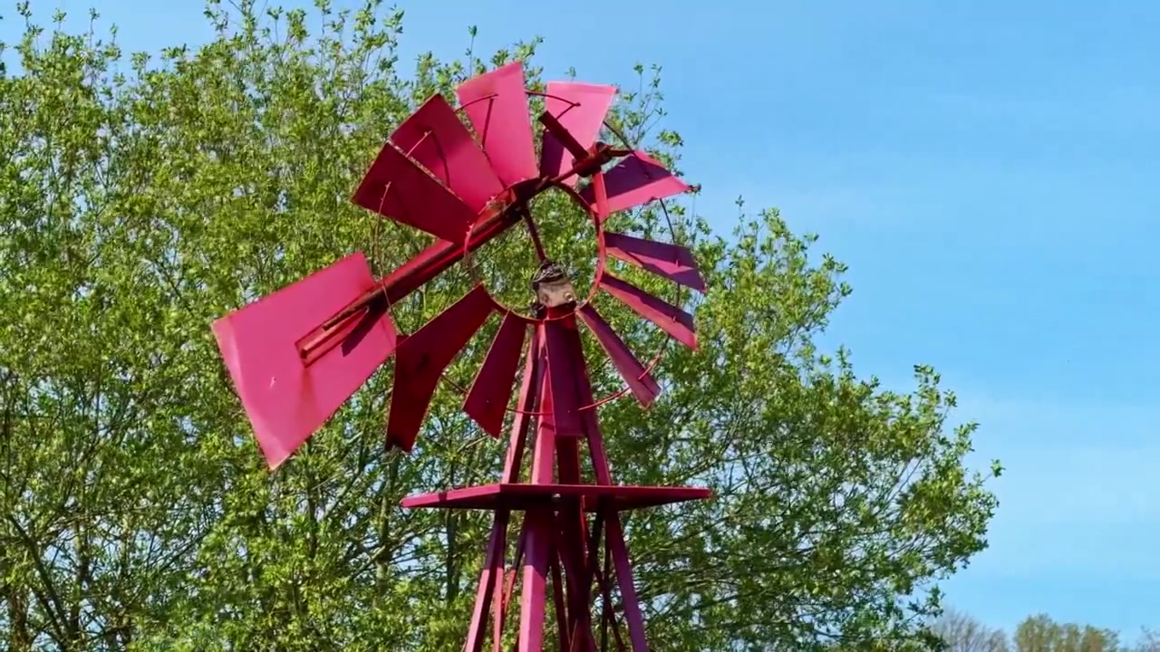 Mulino a vento decorativo in metallo rosso con alberi verdi e cielo blu sullo sfondo.