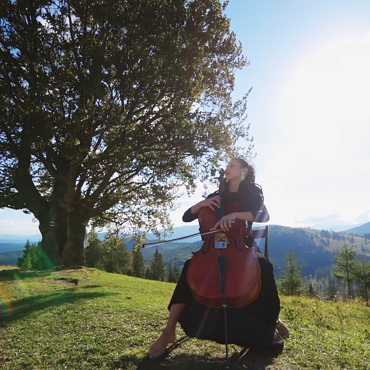 Female music player sitting on the chair plays violoncello. Classical music player at backdrop of mountains on sunny day
