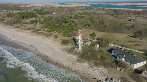 Aerial-establishing-view-of-white-colored-Pape-lighthouse,-Baltic-sea-coastline,-Latvia,-white-sand-beach,-large-waves-crashing,-sunny-day-with-clouds,-wide-drone-shot-moving-backward,-camera-tilt-up