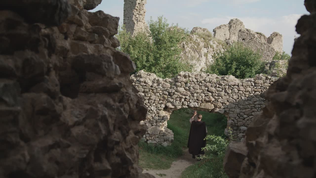 Viewed through a gap in a stone wall, a man in a historical brown cloak walks along a path and through an ancient archway in the ruins of a medieval fortress.