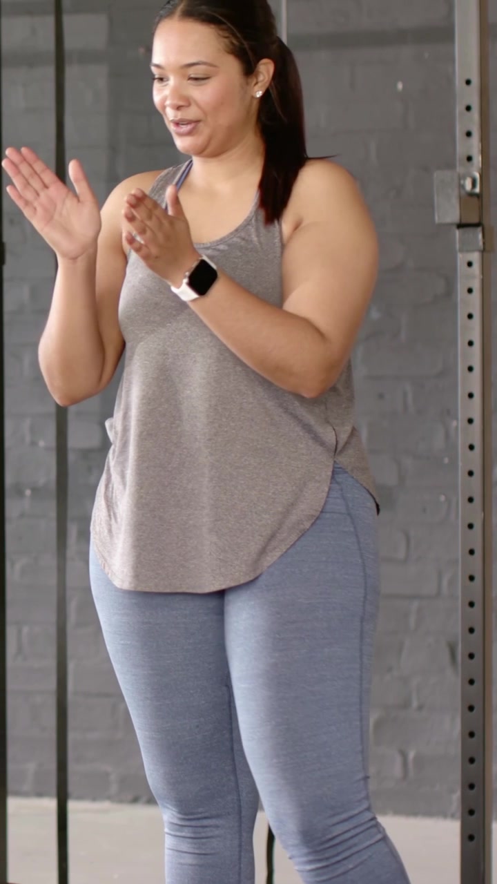 Video verticale: Donna che applaude in canotta e leggings, che allena e gesticola a destra presso lo squat rack della palestra.