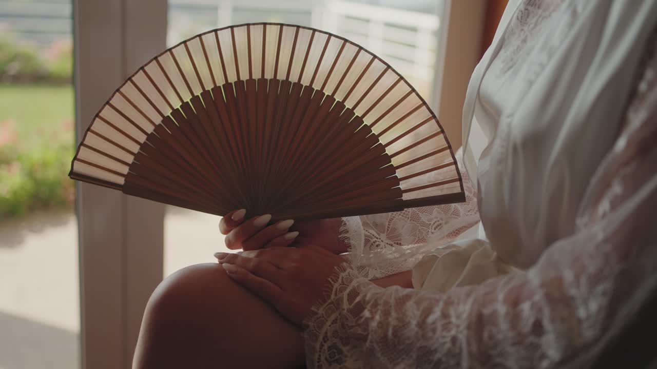 Close up of bride holding wooden hand fan with lace robe and long nails in natural light