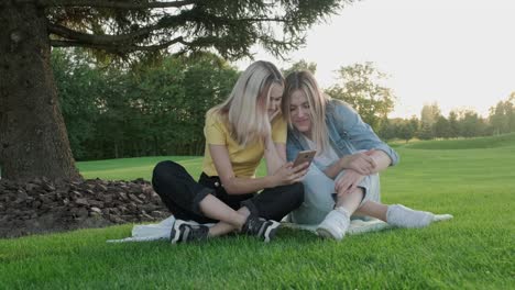 two girlfriends sitting on green grass of lawn smiling talking looking together at smartphone screen