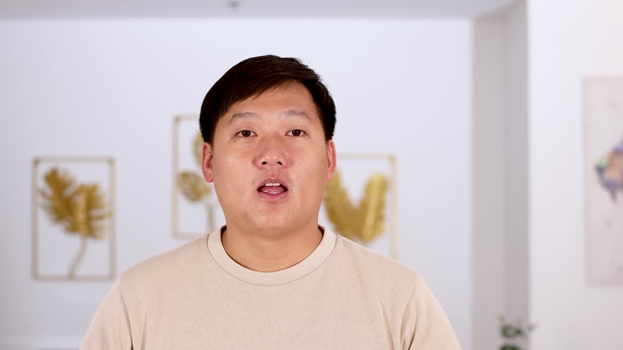 Asian man reacts with a surprised facial expression, covering his mouth with his hands in a bright, modern indoor setting with soft lighting and minimal camera movement
