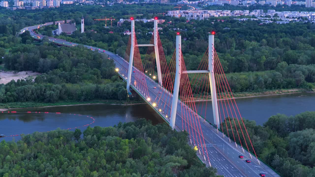 Lighting Siekierkowski bridge over Vistula River and driving cars in Warsaw, Poland. Dusk scene in the evening. Suburb blocks in background. Low housing area of polish town. Aerial wide shot