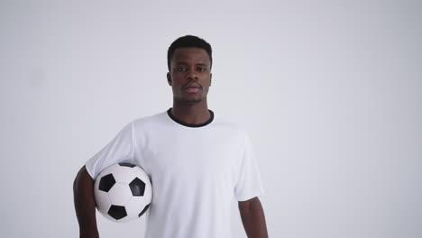 retrato de un futbolista étnico africano en un uniforme blanco sobre un fondo blanco caminando con una pelota en las manos mirando a la cámara la cámara se mueve hacia atrás desde el headshot al plan principal