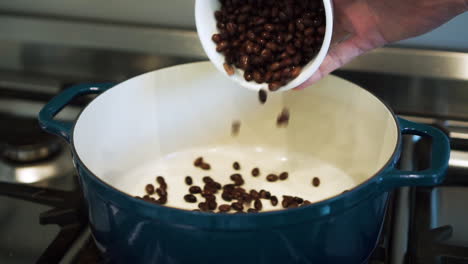 Male-hand-tips-black-beans-into-large-baking-pot-on-stove,-slow-motion-close-up