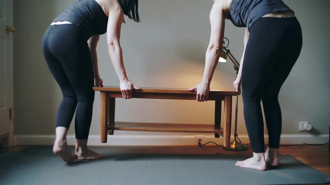 Grasping bench ends, two women in workout clothes lifting to reposition bench near rug and lamp