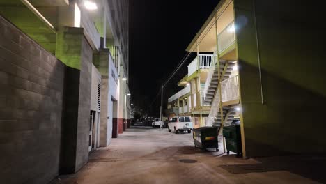 Moving-cinematic-shot-walking-through-an-alleyway-at-night-with-a-towering-building-on-the-left-and-stairs-leading-to-apartments-on-the-right