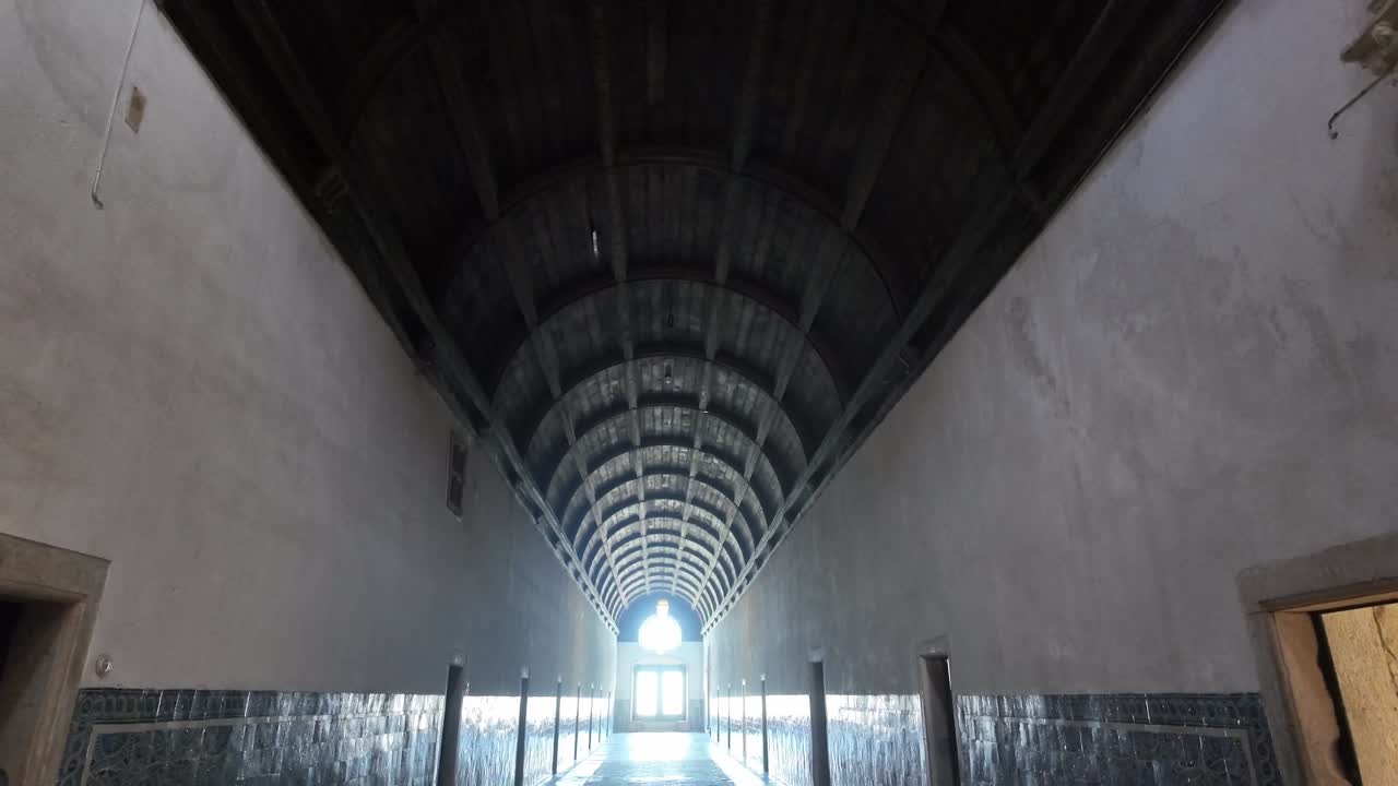 Convent of Christ in Tomar, Portugal, revealing a long cloister corridor with vaulted ceiling, tiled walls, and bright doorway light, handheld tilt upwards view