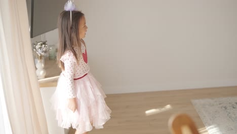 a young girl dances joyfully in a bright, cozy living room. she wears a pink dress with a heart design, moving playfully in the sunlit, minimalist space with neutral decor and soft natural light.