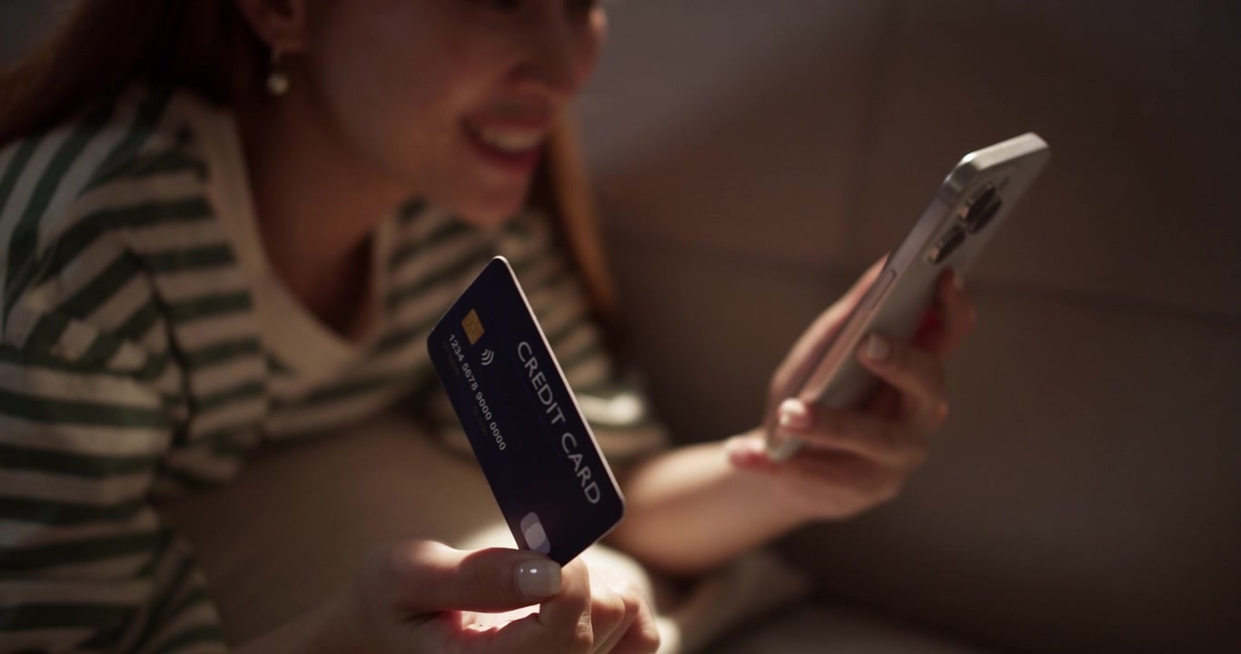 Una giovane donna sorride mentre fa acquisti online, tenendo la sua carta di credito e il telefono, riflettendo la gioia della spesa digitale