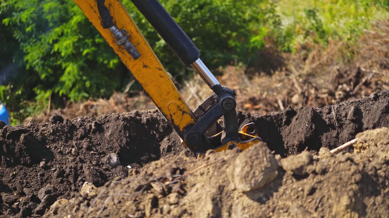 Bucket of tractor digs the ground. Excavator digging the soil on the background of green trees in summer. Close-up.