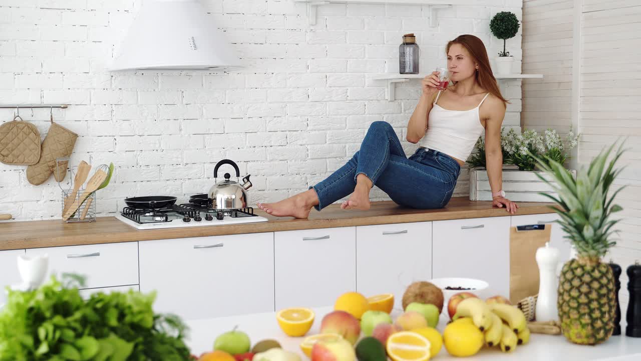 Frullato vegano salutare con frutta. Bella ragazza felice a casa in cucina beve succo fresco da un bicchiere. Tavolo con diversi frutti di stagione.