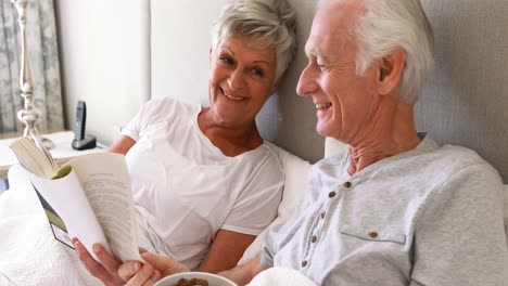 Senior-man-having-breakfast-while-woman-reading-a-book-on-bed