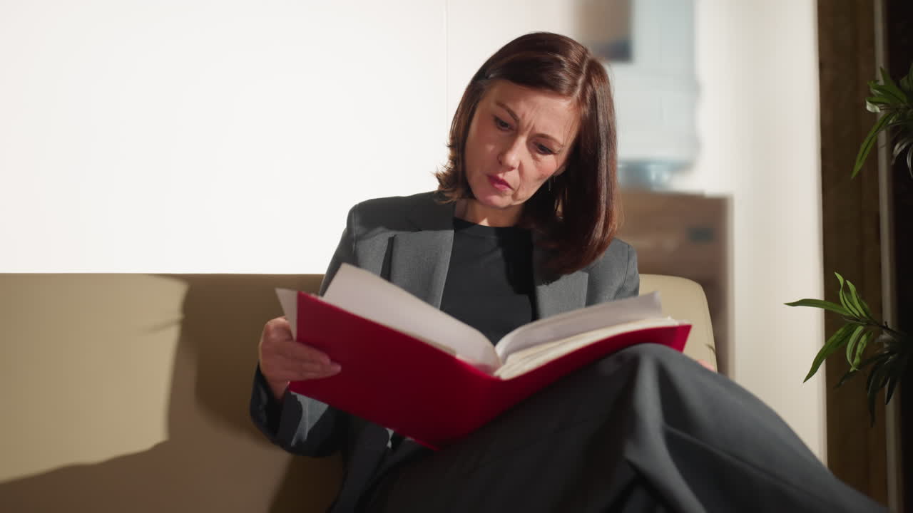 Businesswoman sitting cross-legged on couch carefully reading red folder with focused expression in bright modern office setting, possibly reviewing legal or business documents