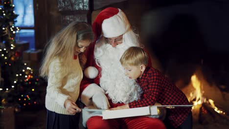 children look into album in santa's hands near fireplace. santa claus with white beard telling stories to little kids at christmas time indoors.