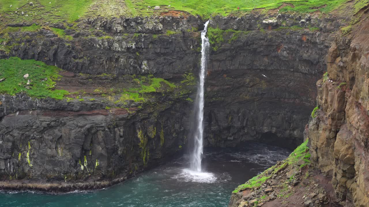 cascada de mulafossur en las islas feroe con una vista lejana de una persona caminando sobre un acantilado