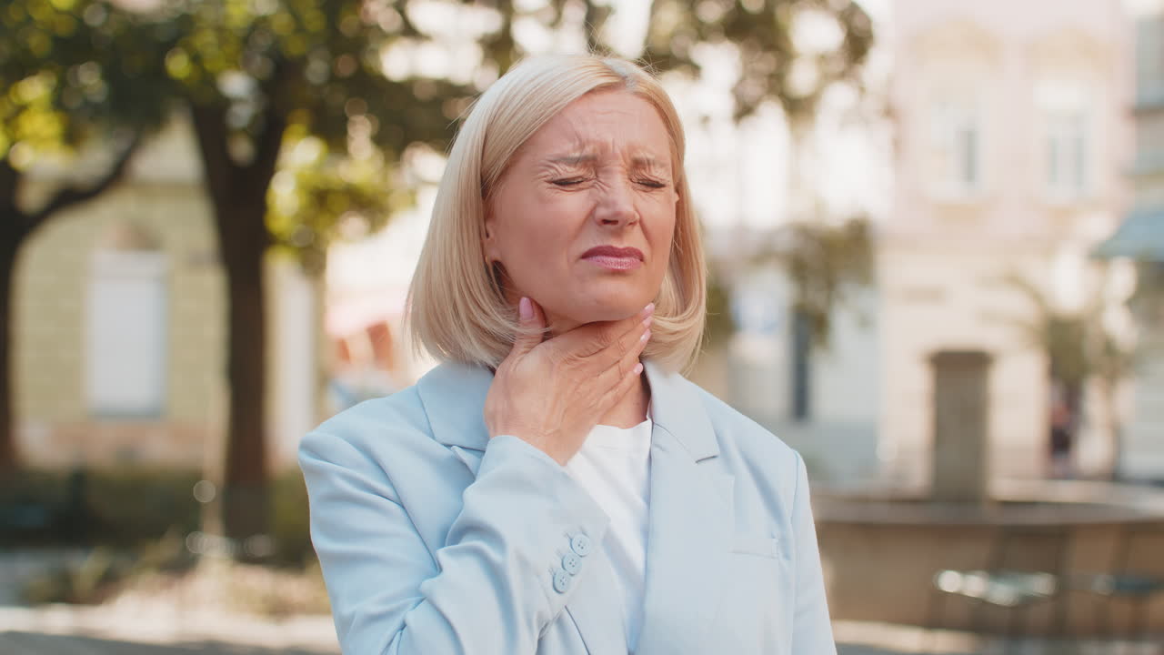Sick mature businesswoman in formal suit suffering from sore throat while standing on city street