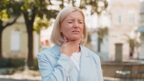 sick mature businesswoman in formal suit suffering from sore throat while standing on city street