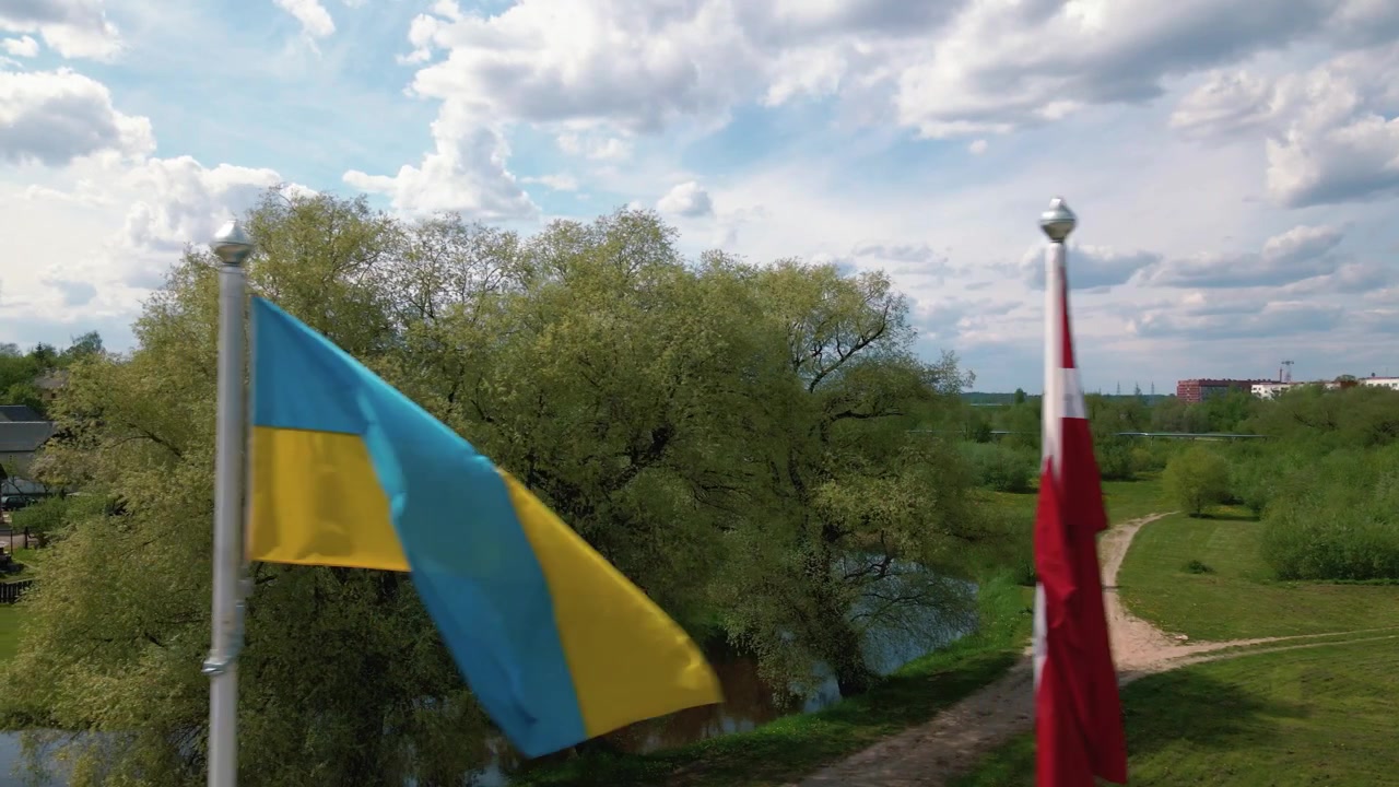 Estas imágenes capturan cinco banderas ondeando suavemente mientras el dron se mueve lateralmente revelando un paisaje vibrante. Las banderas mostradas incluyen las de Ucrania, Letonia, Rezekne y la Unión Europea.