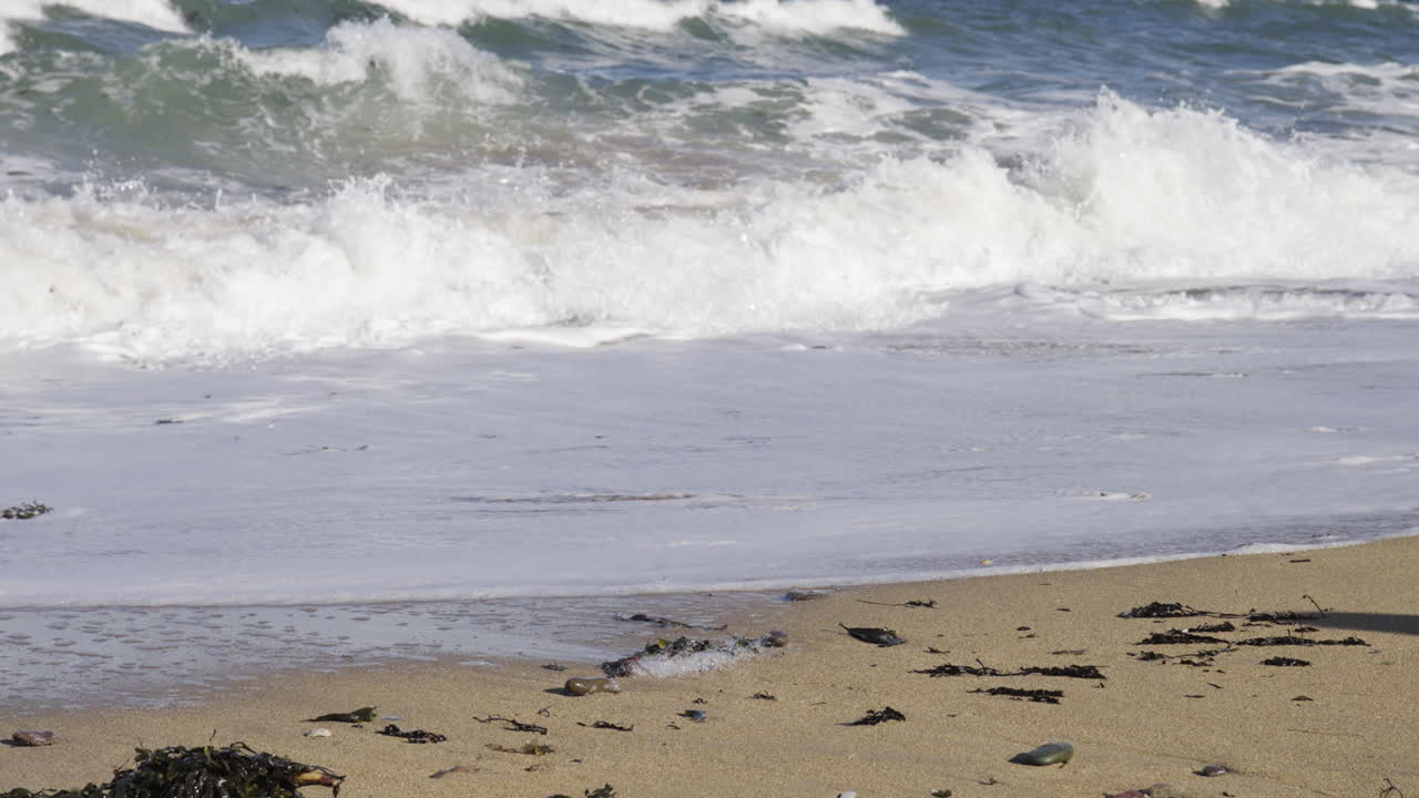 Primo piano cinematografico di schiuma di mare bianca e onde oceaniche blu che si infrangono su una spiaggia di sabbia dorata con alghe. Perfetto per vlog di viaggio, sfondi per la meditazione e concept per le vacanze estive.