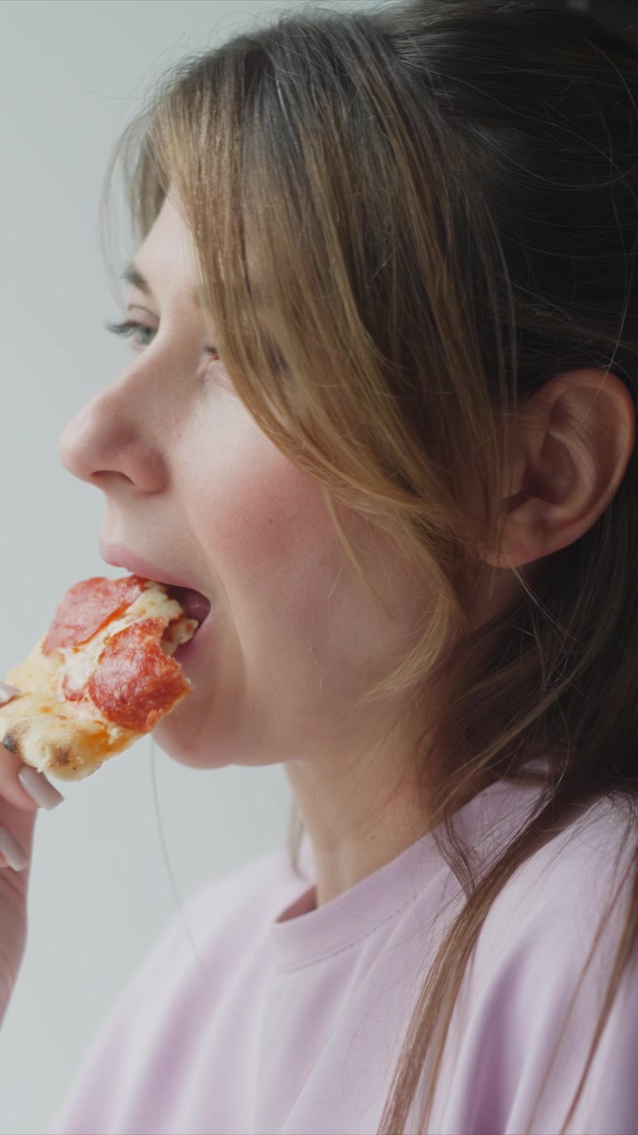 Close up of woman eating pepperoni pizza. Woman holds slice with her hand bringing it to her mouth and taking bites against plain white background. Soft lighting highlights facial features