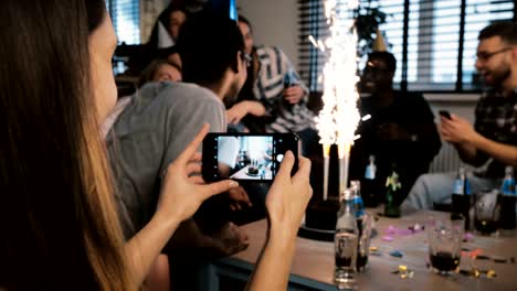 female hands take a smartphone picture of multiethnic birthday party with firework candles, friends hugging slow motion