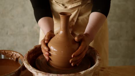 cheerful, young good looking woman wearing apron sitting in pottery and making clay jug on potter wheel using hands