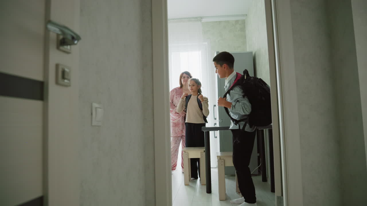 girl and boy wearing casual clothes preparing school bags in morning as mother watches nearby in pajamas, home interior lit by window light, siblings getting ready for school day, backpacks on shoulders