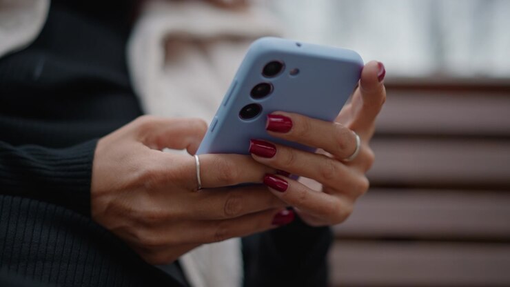 Mani che toccano e scorrono attivamente lo schermo dello smartphone mentre si è seduti su una panchina di legno all'aperto, mostrando unghie rosso scuro, anelli d'argento e una postura casuale e rilassata alla luce soffusa del giorno con sfondo sfocato