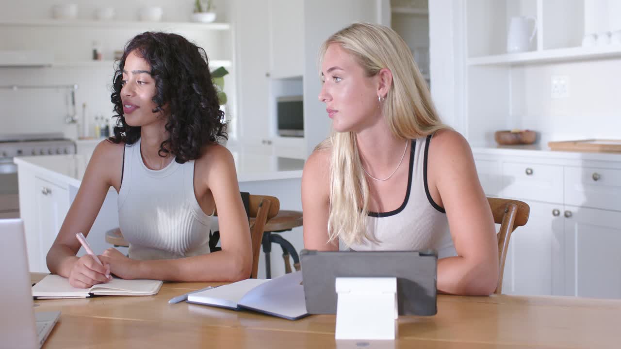 Mujeres diversas colaborando en una mesa en la cocina, la mujer de la derecha proponiendo, la mujer del centro escribiendo en notas.