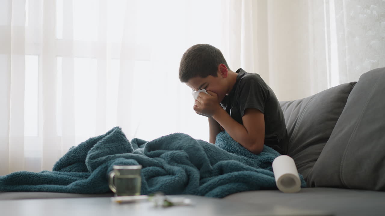 Sick young child sneezing into tissue on couch with blanket covering him, showing tiredness near window with glass cup on table, feeling unwell and needing rest to recover from cold