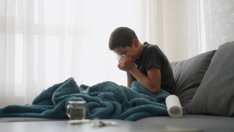 sick young child sneezing into tissue on couch with blanket covering him, showing tiredness near window with glass cup on table, feeling unwell and needing rest to recover from cold