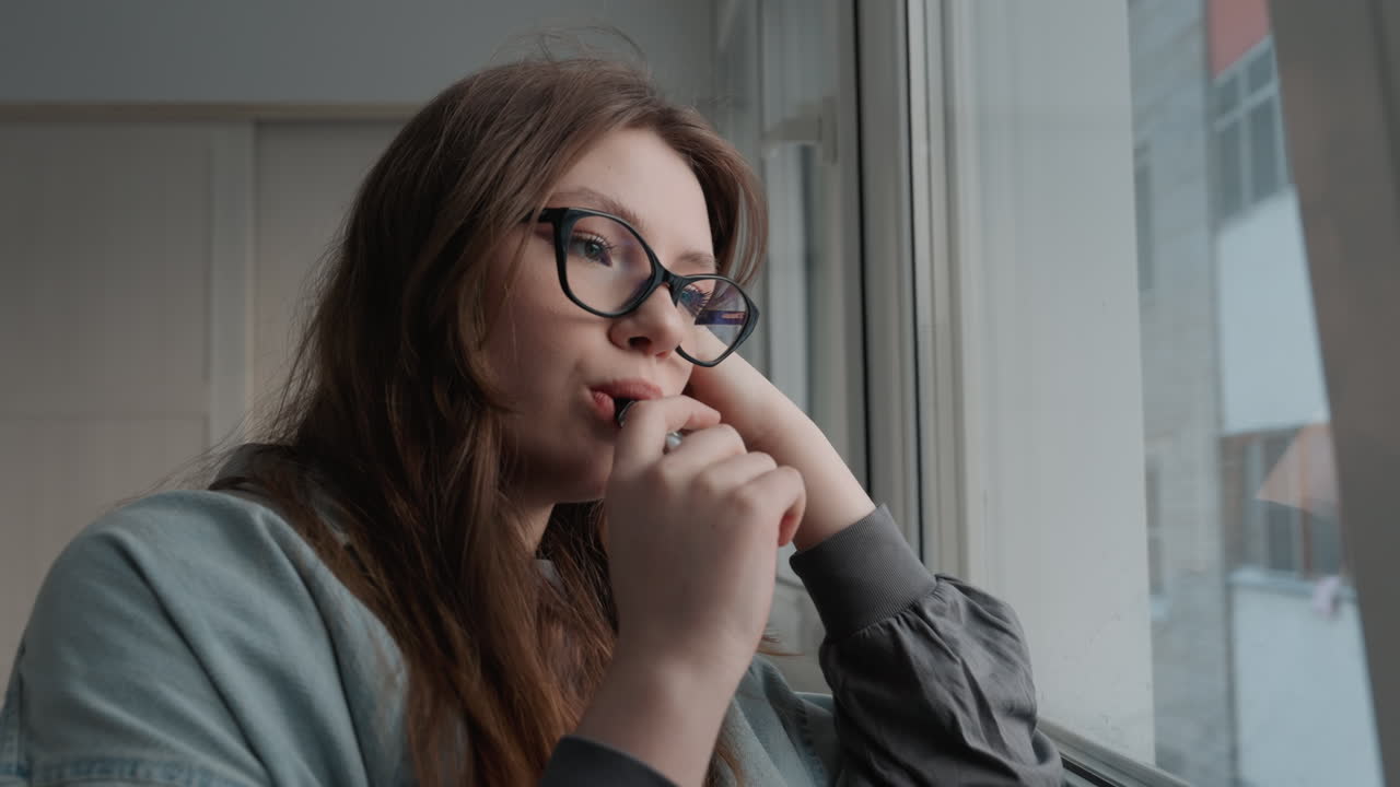 White woman at window holding mug, pensive graduate student in glasses, cozy sweater, morning light through pane, small apartment interior, reflective mood, sipping tea, looking out to urban streets