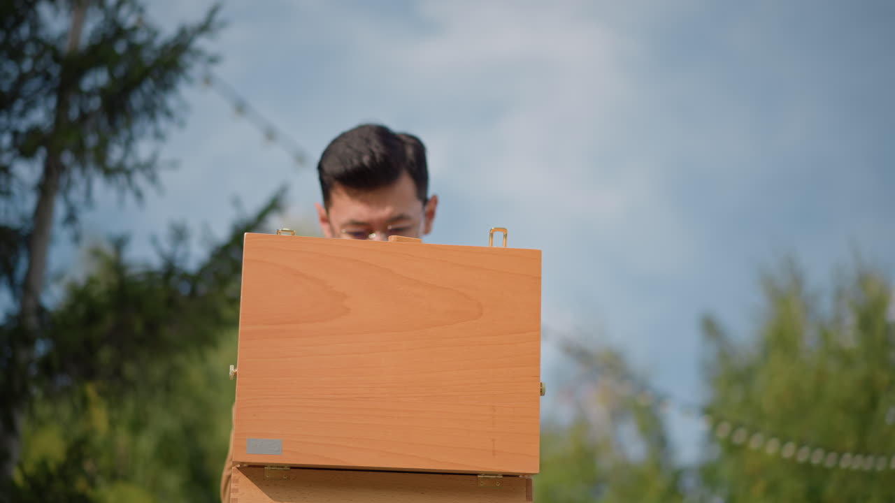 Pria Asia membongkar kotak lukisan kayu, penggambar sketsa jalanan yang tenang sedang mempersiapkan sesi plein air, membuka koper vintage, menata kuas dan kanvas, mempelajari referensi, ekspresi penuh pertimbangan, langit biru