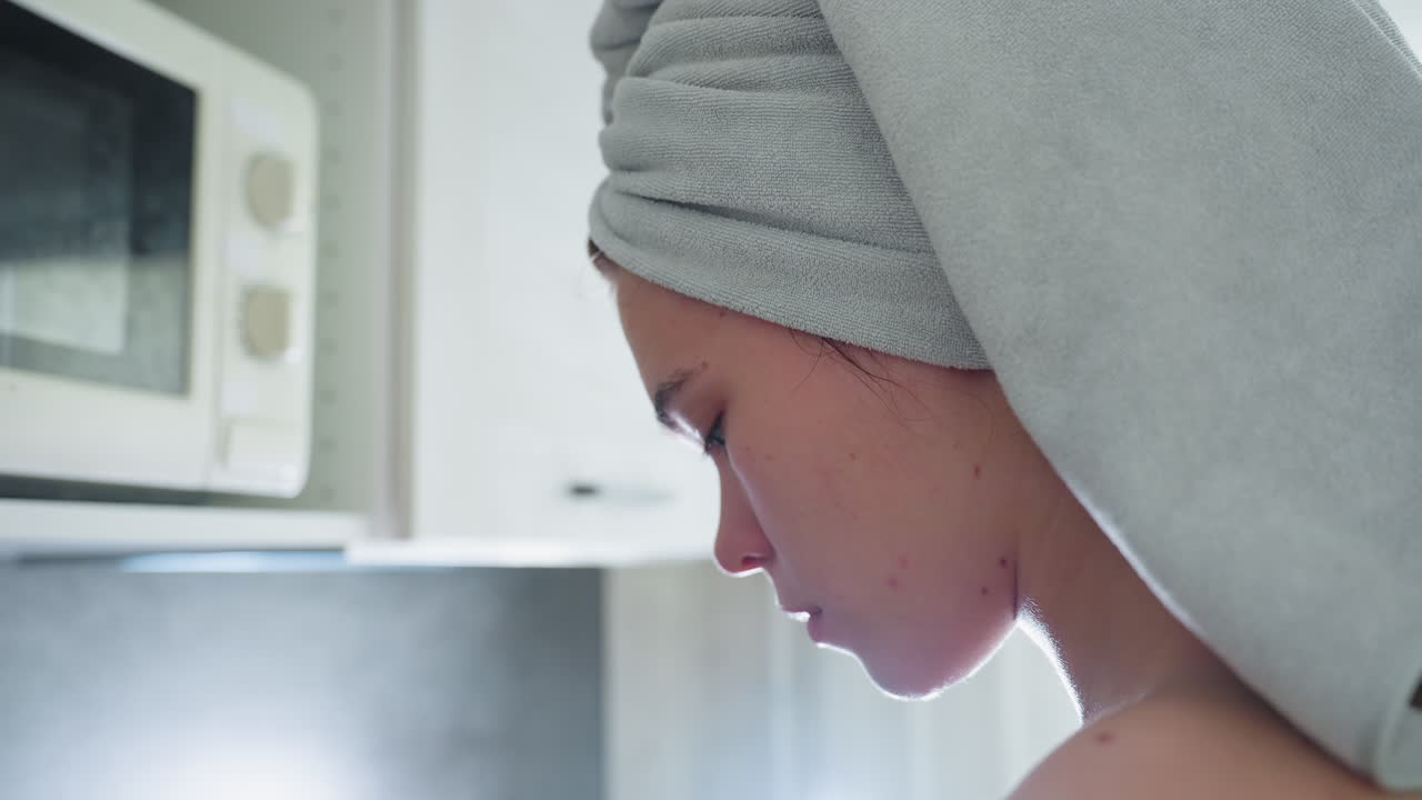 Close-up of woman with towel-wrapped hair, standing in kitchen, focused on chopping cucumber, bright kitchen background with contemporary appliances, capturing a healthy cooking moment