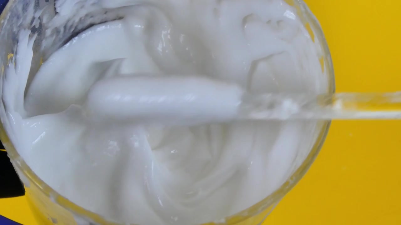 Espátula de laboratorio removiendo una emulsión de crema cosmética blanca en un vaso de precipitados de vidrio sobre un fondo amarillo.