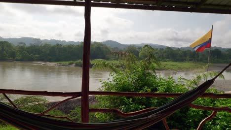 Die-Flagge-Ecuadors-Weht-Im-Wind,-Mit-Einer-Hängematte-Im-Vordergrund-Und-Blick-über-Einen-Balkon-Auf-Den-Amazonas-Regenwald-Gegenüber-Dem-Rio-Napo-In-Ecuador,-Südamerika