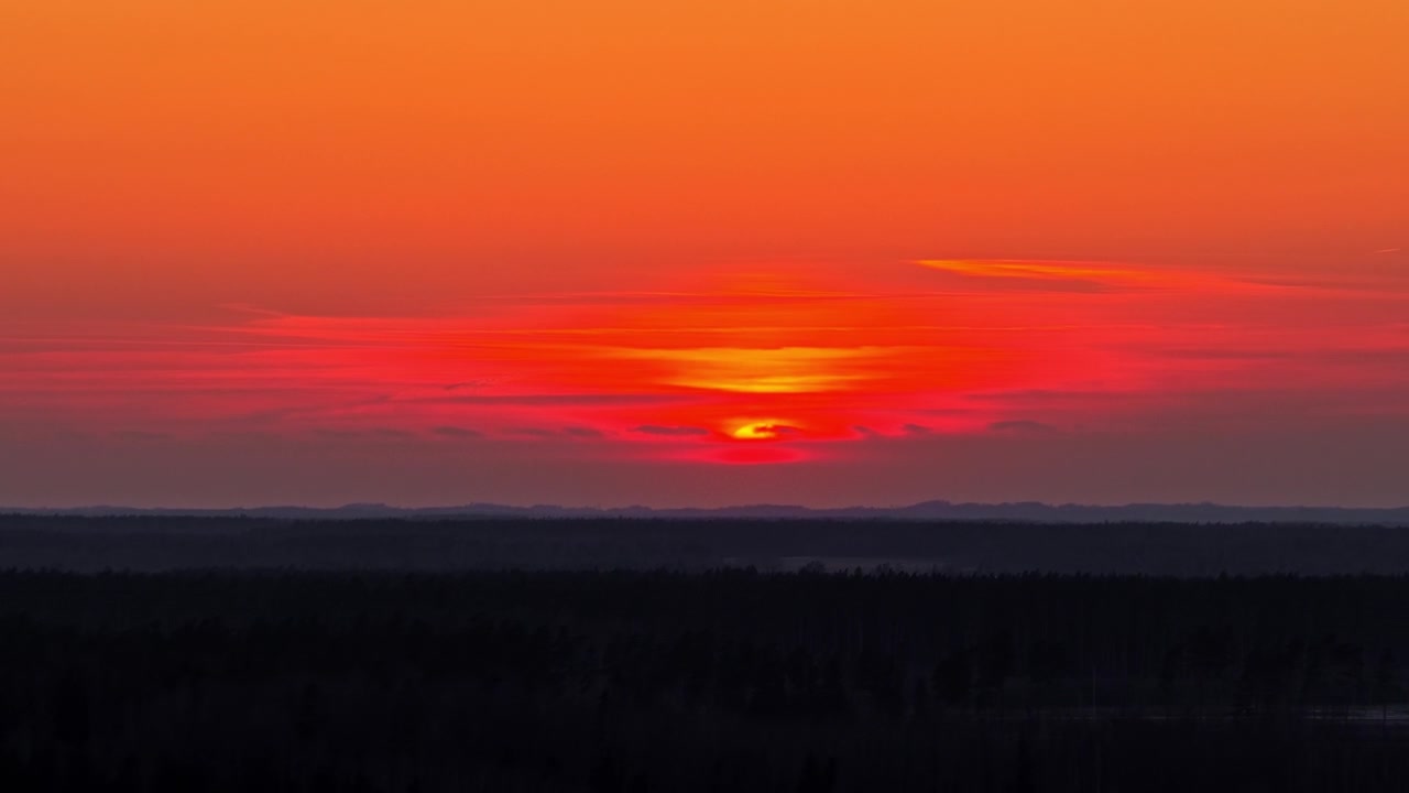Rote Sonne, die über einem dunklen Waldhorizont unter einem dramatischen und farbenprächtigen Wolkenhimmel untergeht.