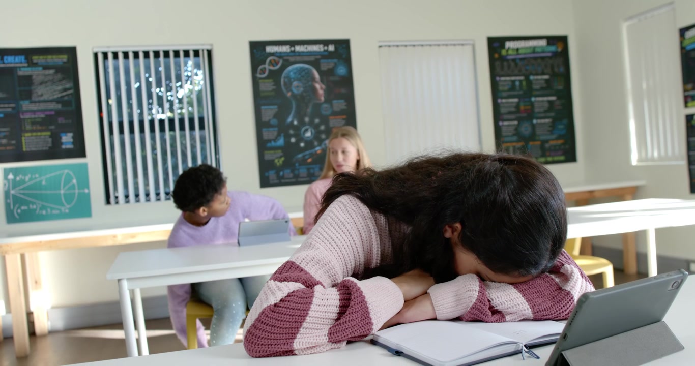 Diverse teenage females in classroom watching front student nodding off onto notebook and tablet