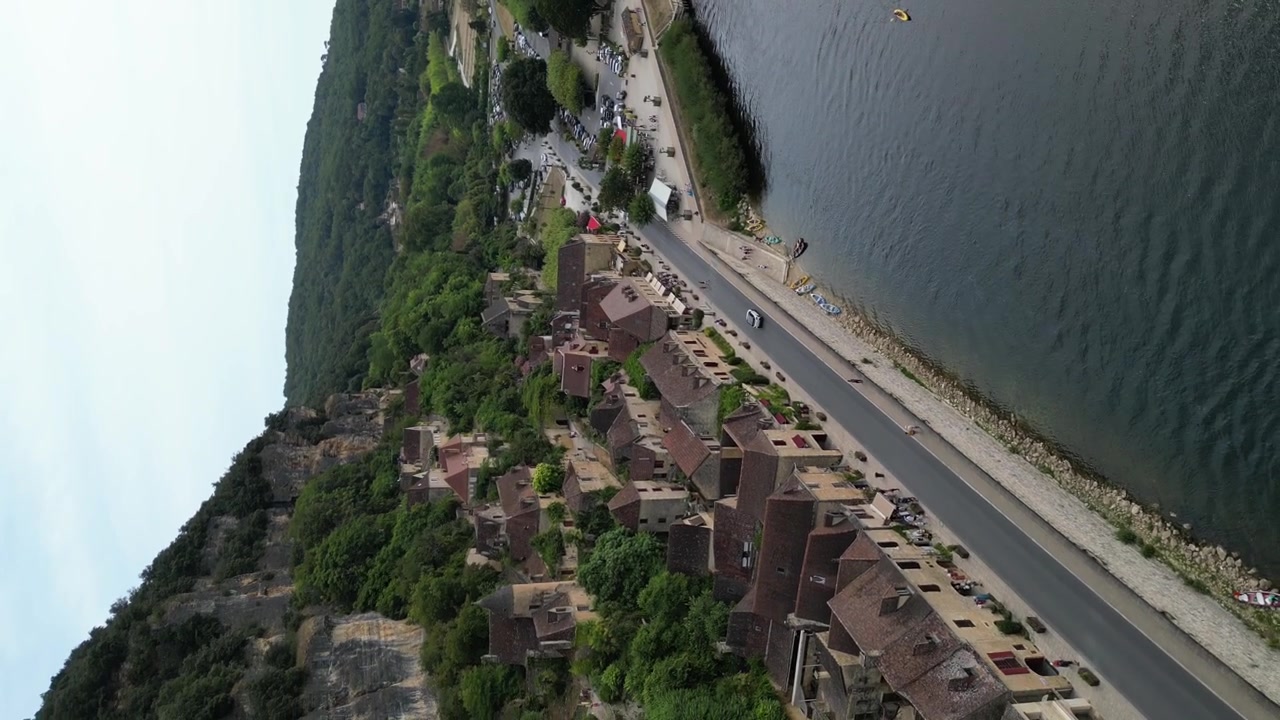 Vista aerea con drone nella campagna francese di una piccola e antica città medievale sotto una falesia di pietra, circondata da una foresta verde e da un fiume che la circonda, a La Roque Gageac, vista verticale.