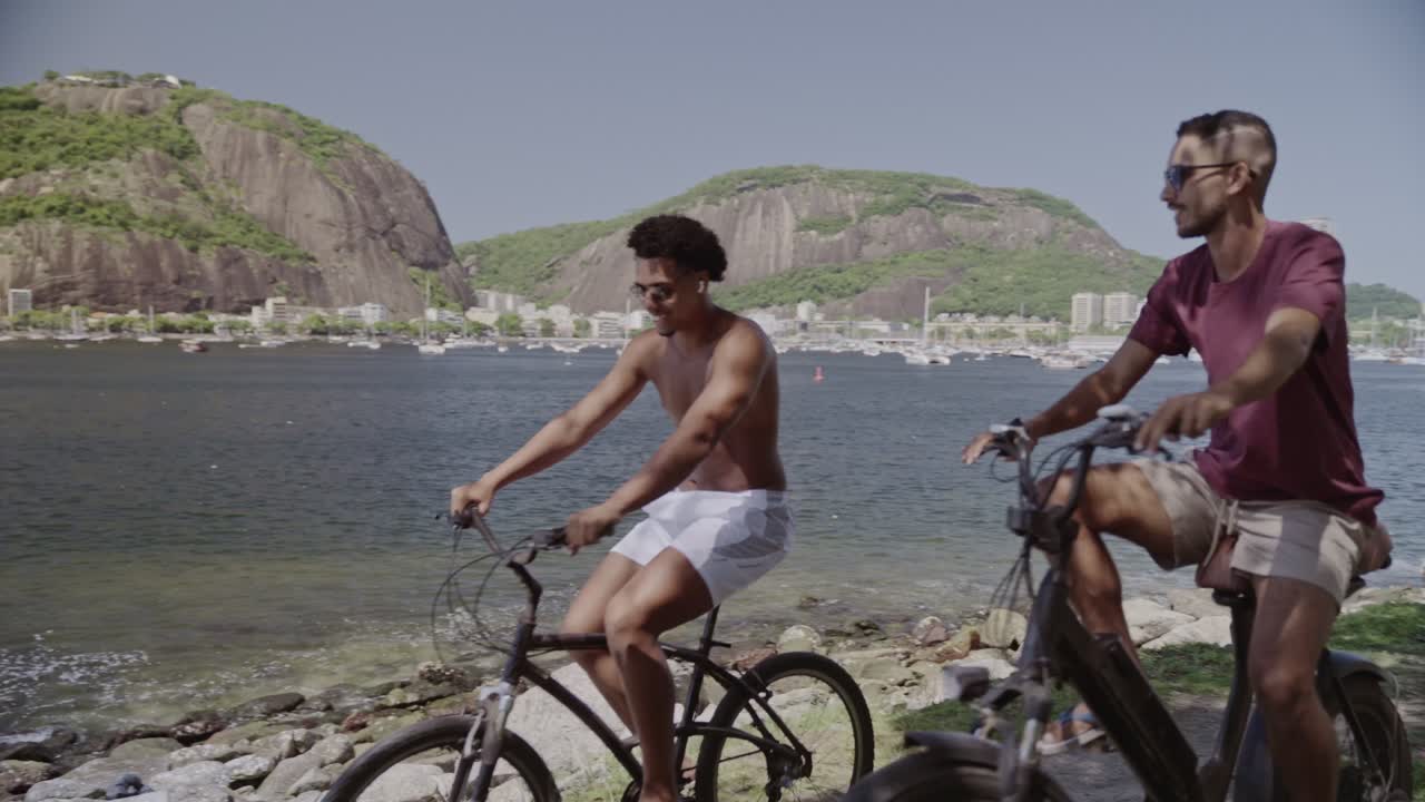Two Men Cycling Along a Scenic Waterfront Path with Mountains and City Views