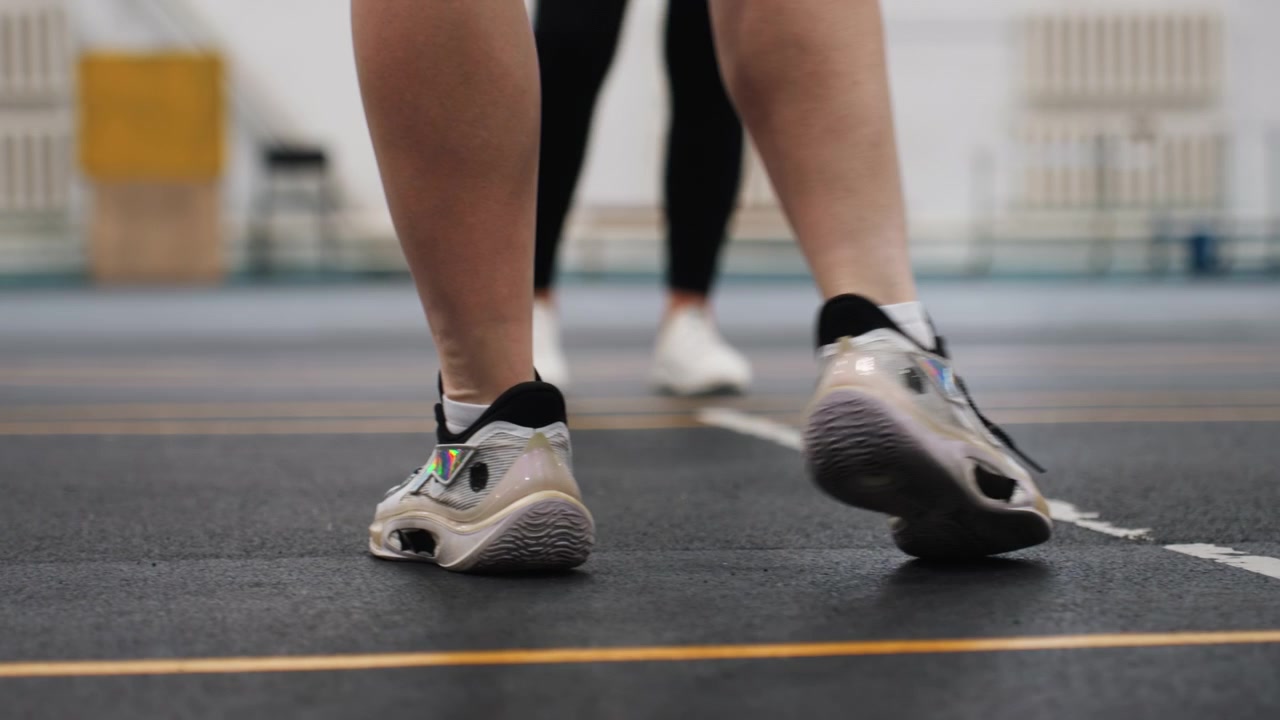Esercizio di gioco di gambe nel badminton su campo al coperto, giocatore concentrato che esegue rapidi passi pivot, dettaglio della suola della scarpa da ginnastica, partner sullo sfondo, superficie del campo in gomma, primo piano dal basso, ritmo costante, intensità del riscaldamento
