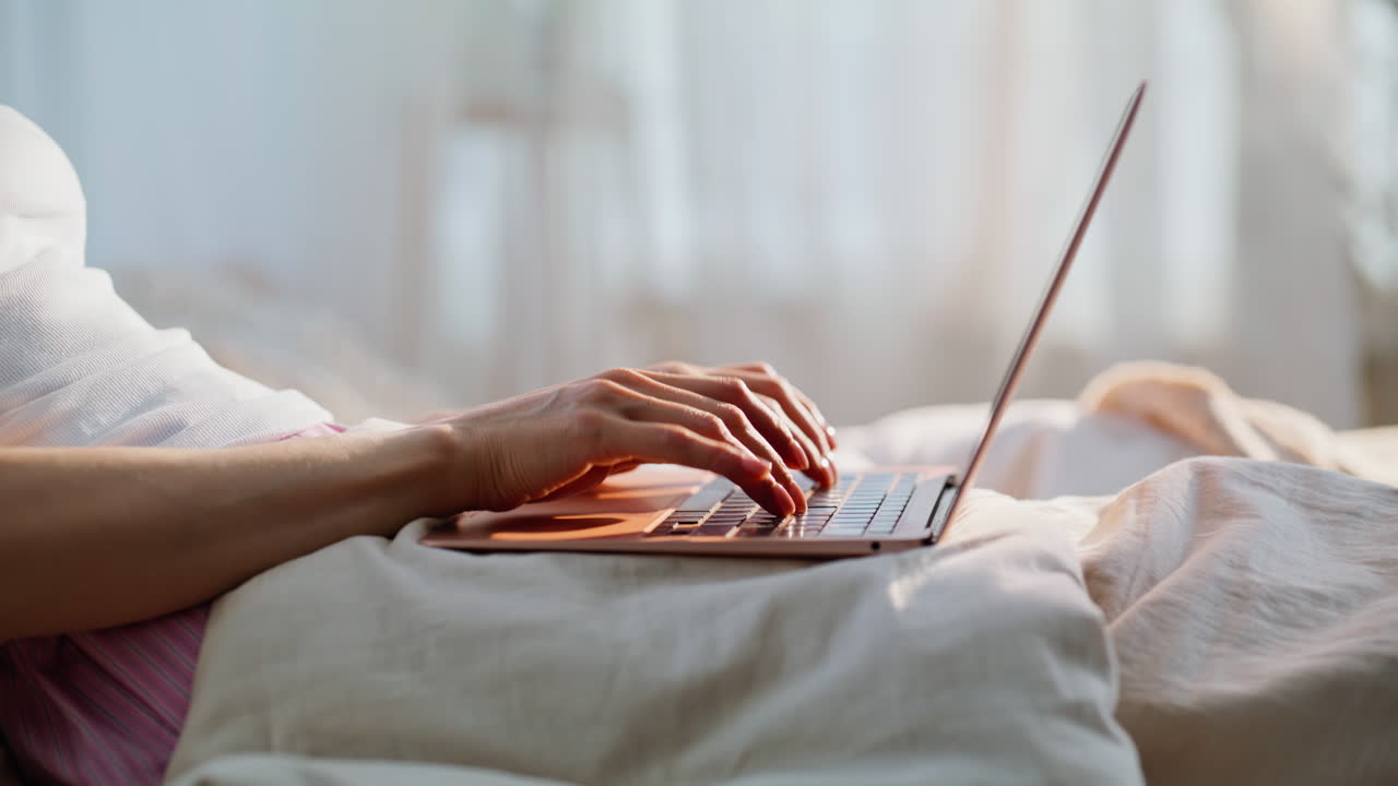 Nærbillede af en freelancers fingre, der skriver på en bærbar computer i en hyggelig seng derhjemme. Uidentificerbar kvinde