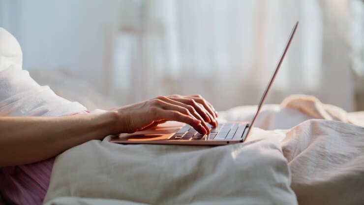 Nahaufnahme von den Händen einer Freelancerin, die auf einem Laptop im gemütlichen Bett zu Hause tippt. Nicht erkennbare Frau