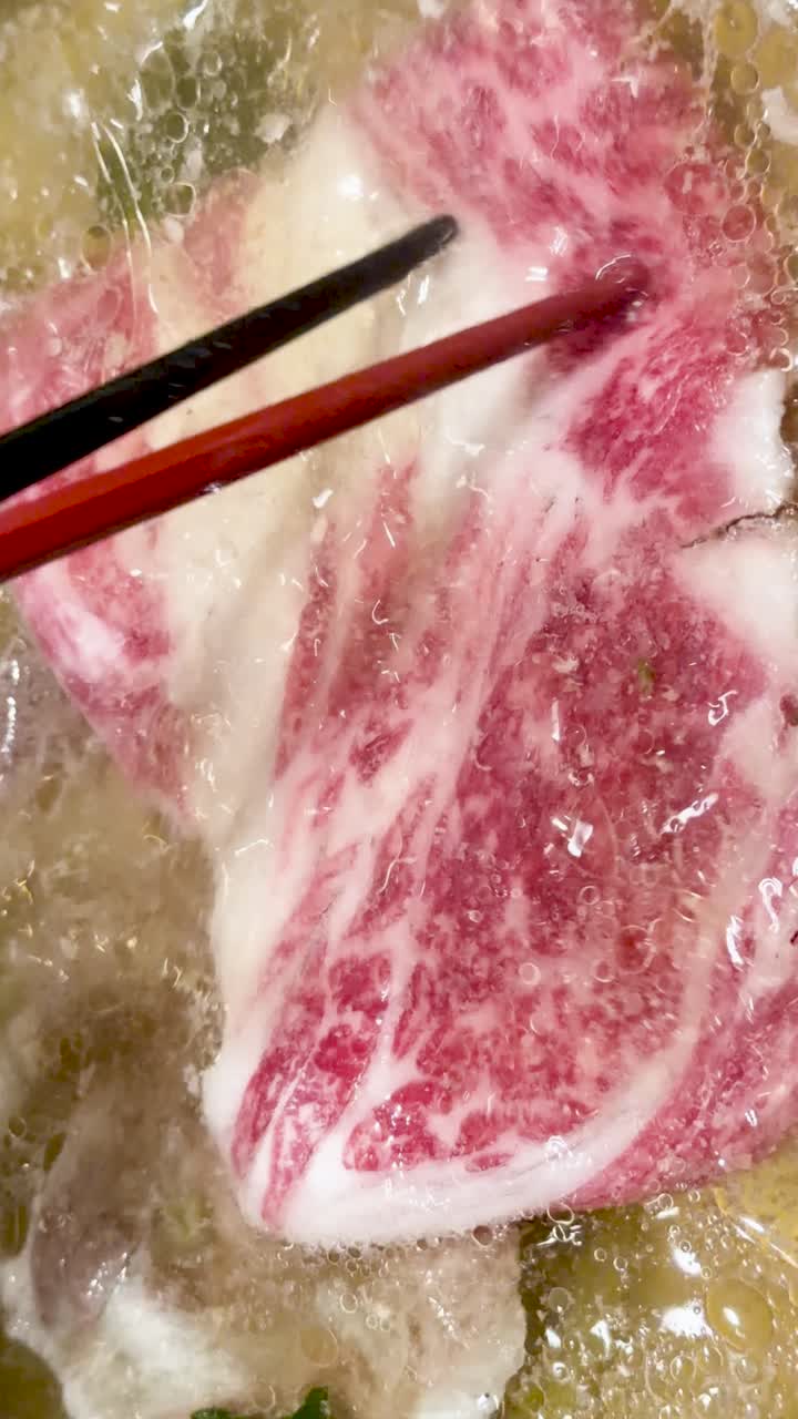 Fettine di manzo Wagyu marmorizzato cuociono in brodo bollente, primo piano, con le bacchette, illuminazione dall'alto.