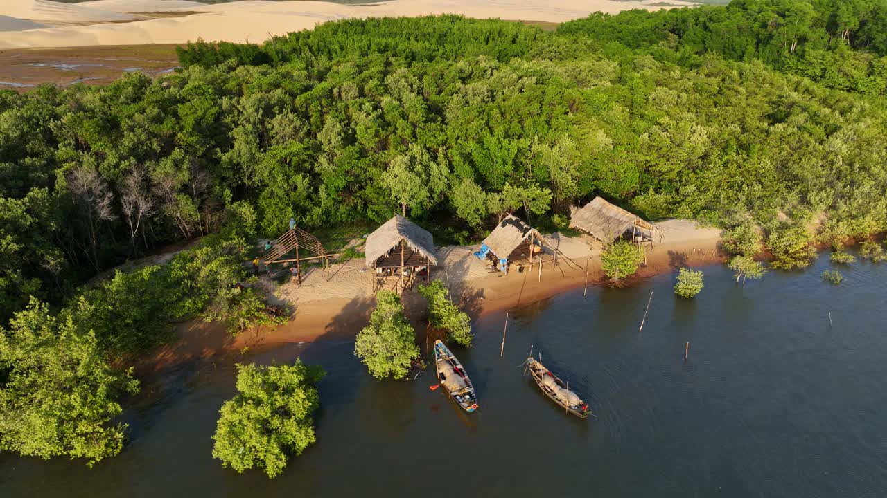 Indigenous people wooden huts near Parnaiba river tributary surrounding mangrove forest, wooden huts for housing near river shore, desert in background, Drone shot
